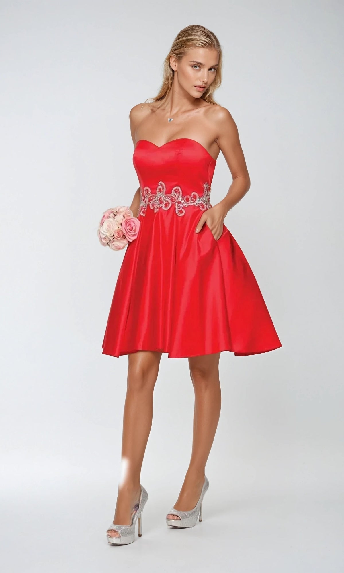 A woman in the Short Hoco Dress: Juliet 769—strapless with a sweetheart neckline and floral belt detail—stands in silver high heels, holding a small bouquet of roses against a plain background.